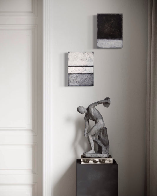 Two black and white paintings against white wall with statue of athlete on stand